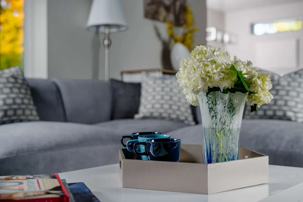 A bright, inviting living space featuring a cozy sofa with patterned cushions, a vase of fresh flowers, and two blue coffee cups on the table. Sunlight fills the room, creating a welcoming atmosphere perfect for relaxing or gathering with colleagues.