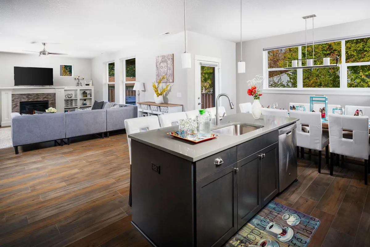 This image captures the open floor plan, featuring the kitchen island, dining table, and living area couch, all visible in a single inviting space. A cozy fireplace anchors the scene, suggesting a central gathering point for a variety of group activities.