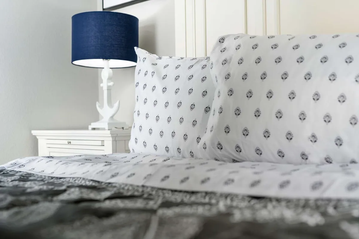A bedroom featuring a bed with inviting patterned bedding, a blue lamp on a nightstand, and a cozy ambiance perfect for reading or relaxing. The soft lighting and tidy setup evoke comfort and tranquility, ideal for winding down after a lively gathering.